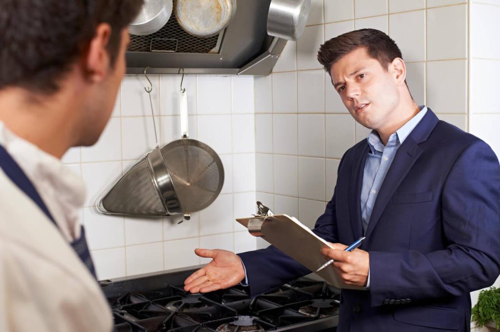 A food inspector with a clipboard discussing requirements with a chef in a professional restaurant kitchen.