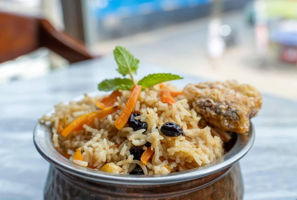 A dish of Halal Afghan Kabuli Pulao, featuring rice mixed with carrots and raisins, served in a traditional metal bowl.